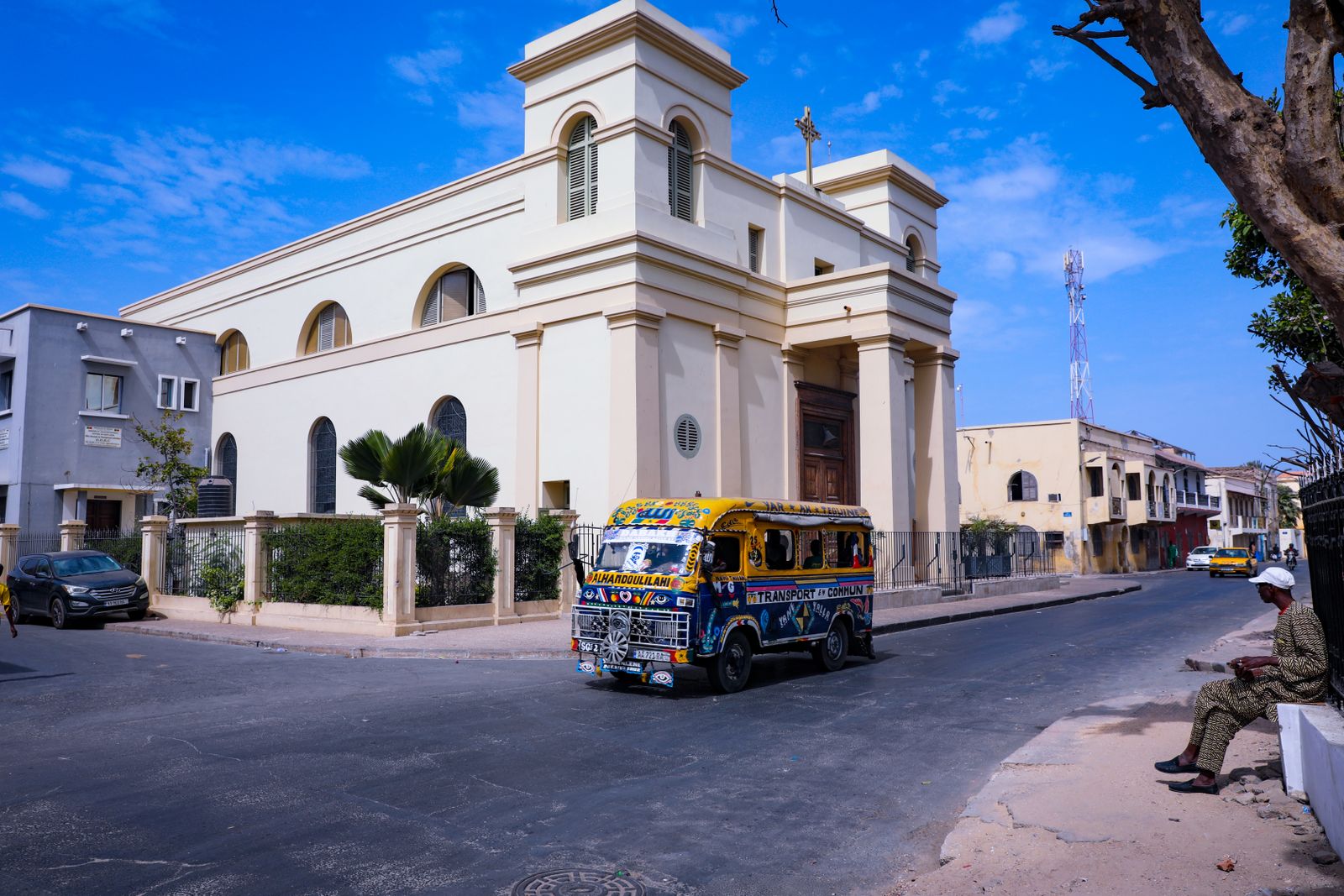 A vibrant minibus travels the streets of Dakar, passing a historic church under a clear blue sky.